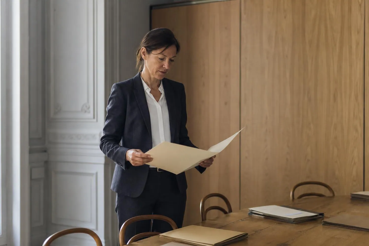 Commissaire aux comptes debout dans la salle de réunion d’un cabinet, un dossier de mandat ouvert à la main, devant une cloison de bois clair
